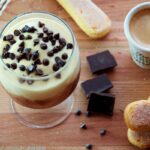 Un bicchiere con del tiramisù coperto con delle gocce di cioccolato. Sul tavolo di legno su cui poggia, dei savoiardi, una tazzina di caffè e qualche pezzetto di cioccolato.